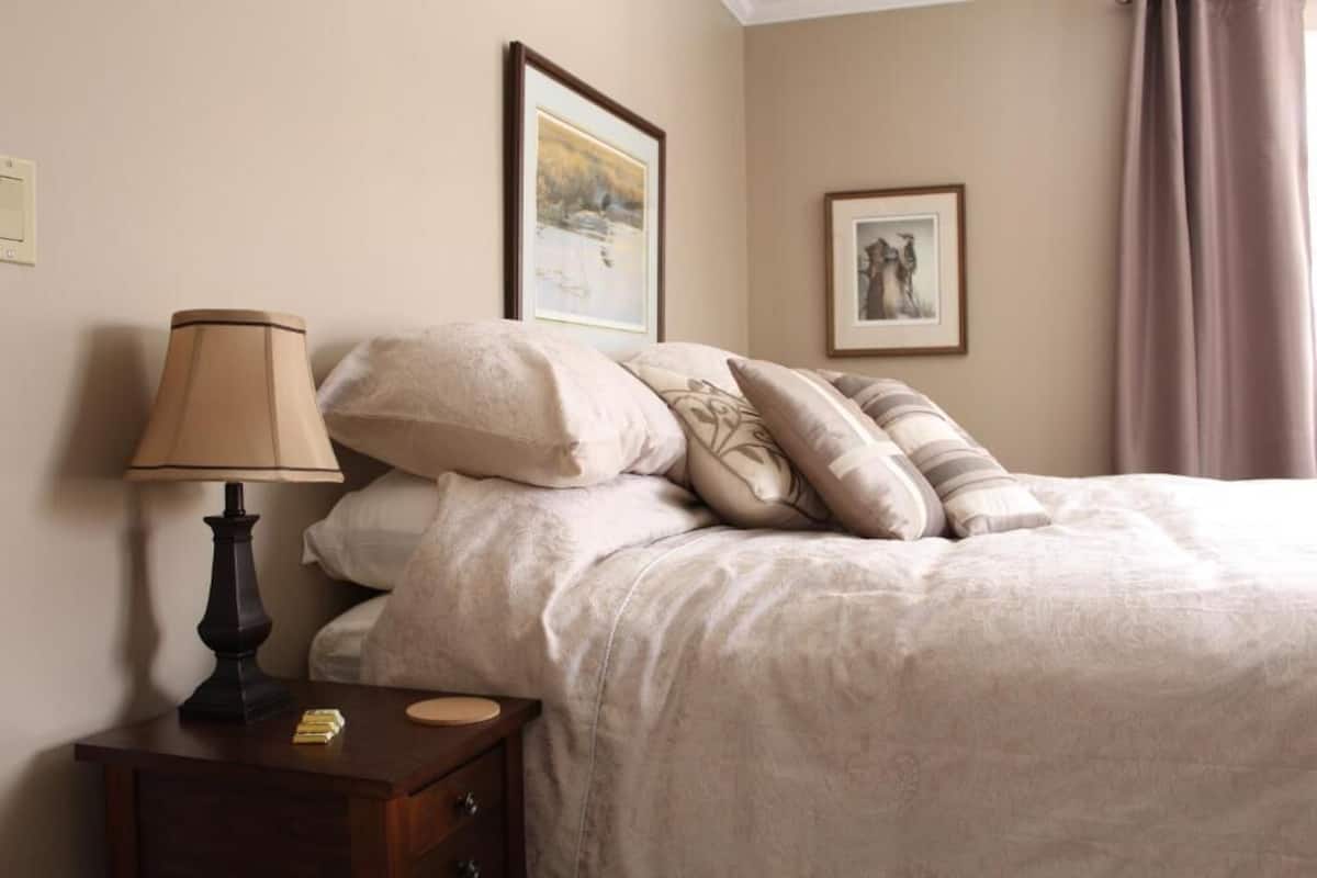 Una cama bien hecha con un edredón color beige y almohadas, una lámpara de noche, y una mesita de noche de madera con una lámpara y algunos objetos encima.