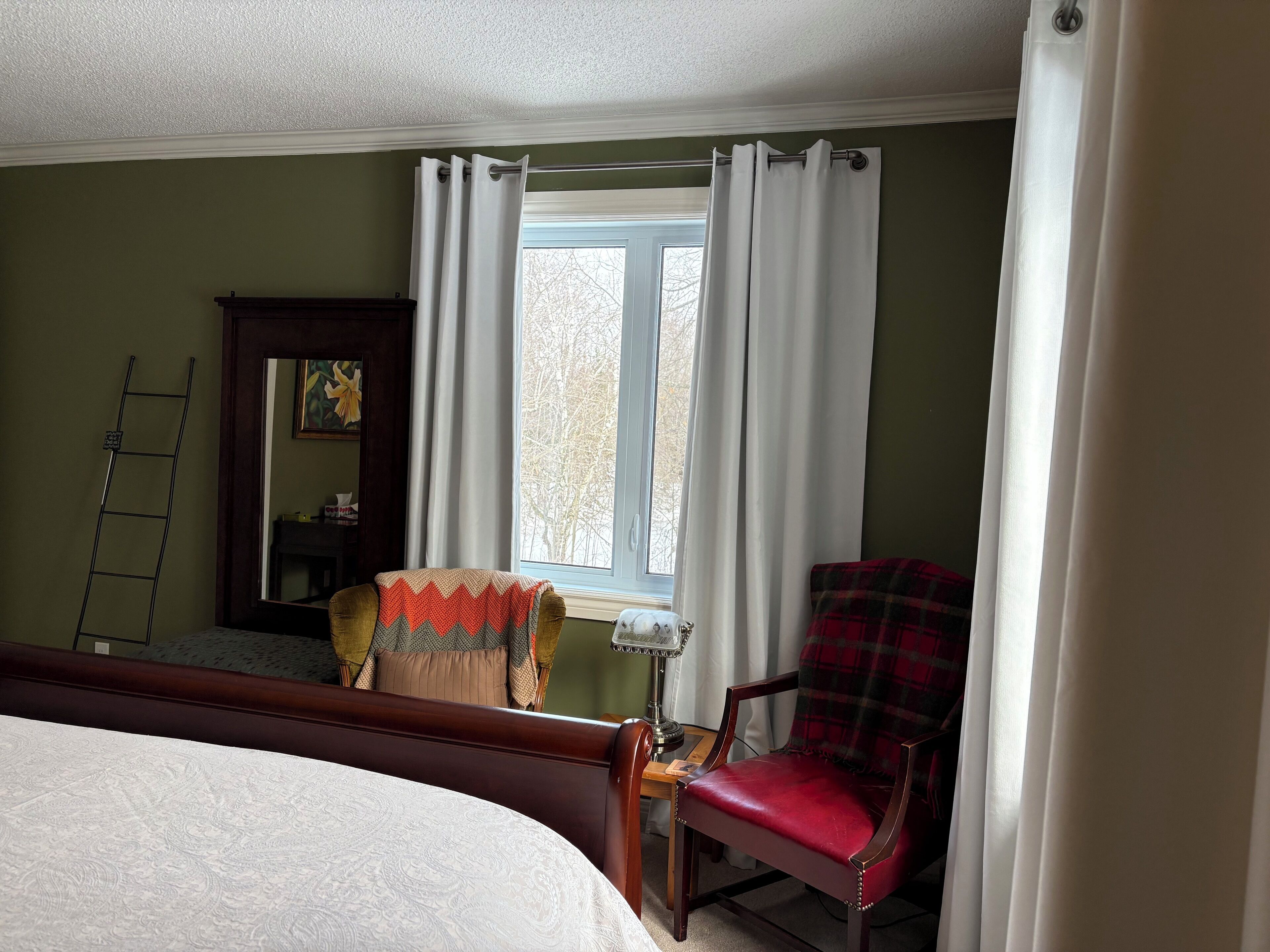 Un dormitorio con una cama, una silla, un ventanal con cortinas, una escalera y una lámpara.