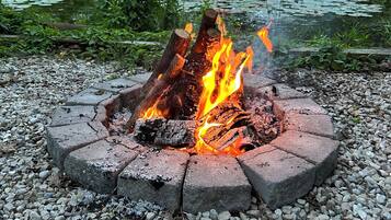 Un hoyo para fuego circular hecho de bloques de piedra con fuego encendido en su interior, rodeado de grava, situado en un entorno natural con árboles y un estanque al fondo.