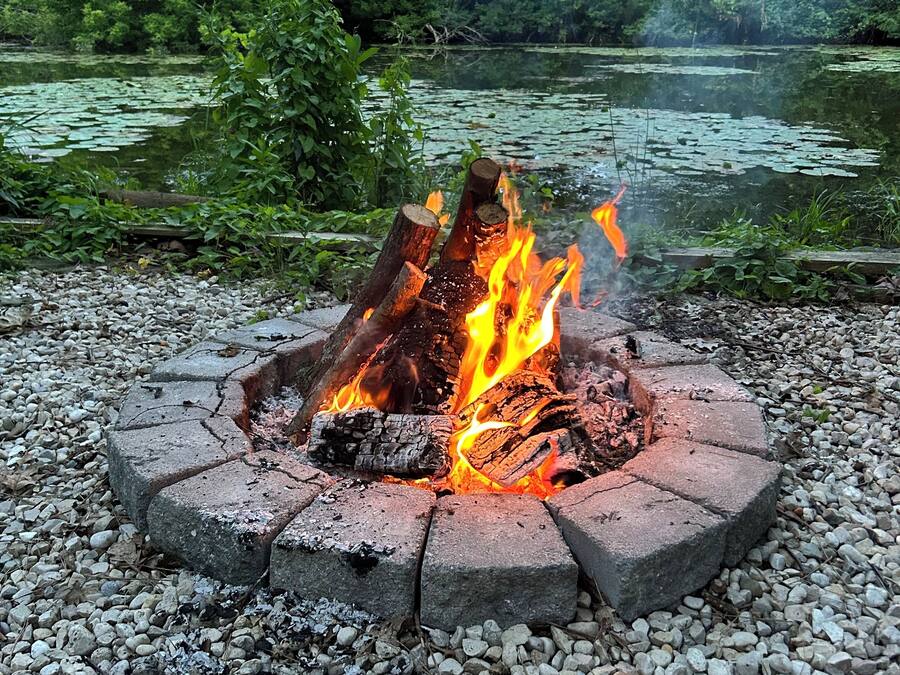 Een ronde vuurkuil van stenen blokken met een vuur erin, omringd door grind, gelegen in een natuurlijke omgeving met bomen en een vijver op de achtergrond.