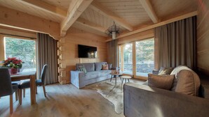 L’intérieur d’un chalet accueillant, avec un plafond en bois, un canapé gris, une table à manger avec des chaises et une grande fenêtre agrémentée de rideaux.