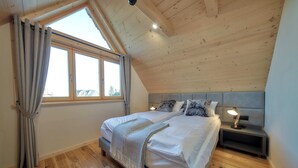 Une chambre avec un plafond en pente, une grande fenêtre avec des rideaux, un lit avec une tête de lit, des tables de chevet avec des lampes et un plancher en bois.