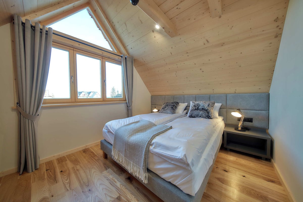 Un dormitorio con techo inclinado, un ventanal con cortinas, una cama con cabecero, mesitas de noche con lámparas y suelo de madera.