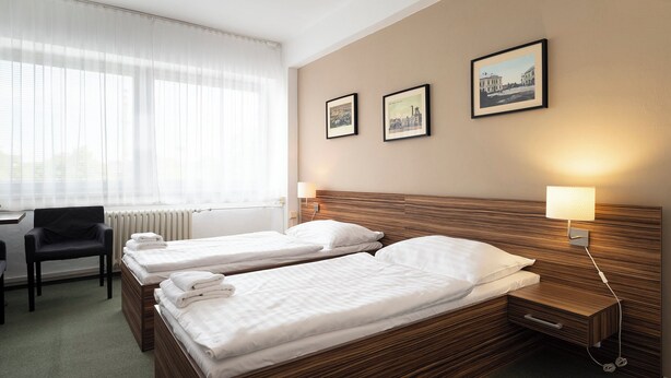 A hotel room with two single beds, a desk, a chair, and a window with sheer curtains.