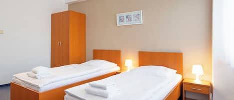 A hotel room with two single beds, wooden headboards, a nightstand with a lamp, and a wardrobe.