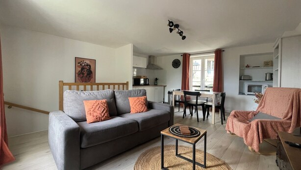 A cozy living room with a grey sofa, a small wooden coffee table, and a pink armchair. A kitchen with a dining table and chairs is visible in the background.