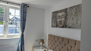 Un dormitorio con ventana, cama con cabecera capitonada, mesita de noche con lámpara y una obra de arte colgada en la pared.
