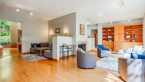 Un salón amplio con suelo de madera, un sofá gris, un sillón azul y una estantería de madera llena de libros.