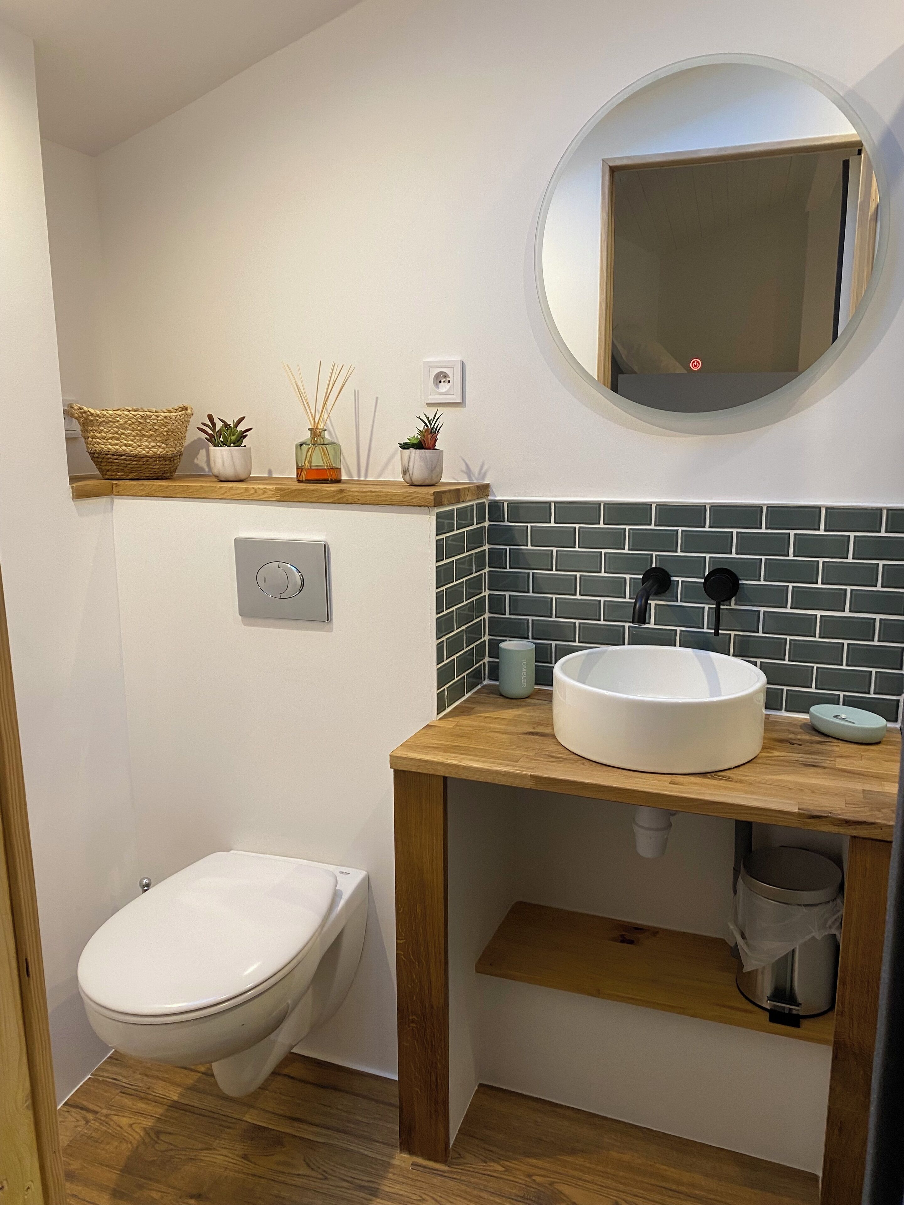 A compact bathroom with a white toilet, a wooden vanity with a round mirror, and a green-tiled backsplash.