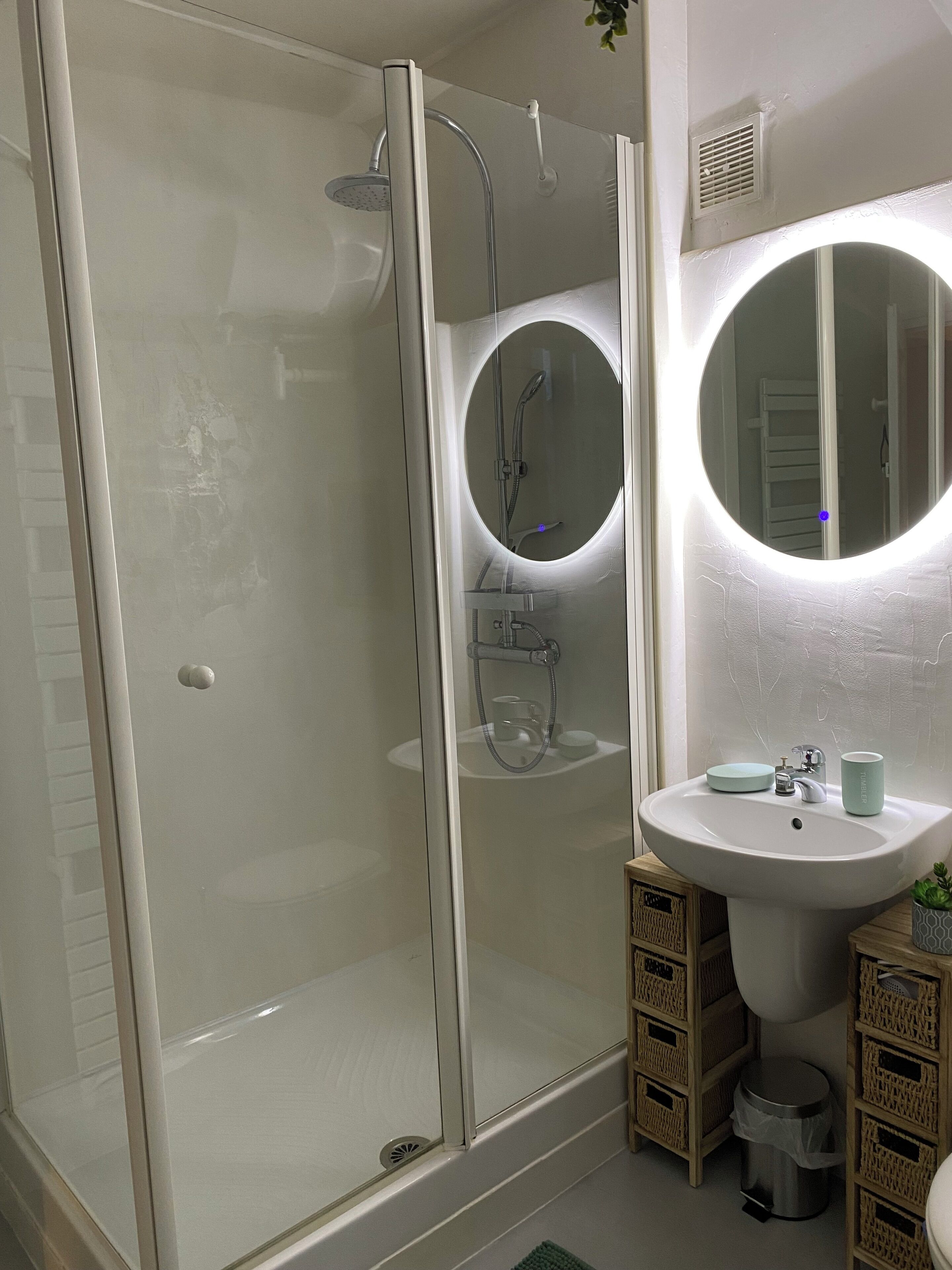 A modern bathroom with a glass-enclosed shower, a white sink with a round mirror, and wicker baskets.