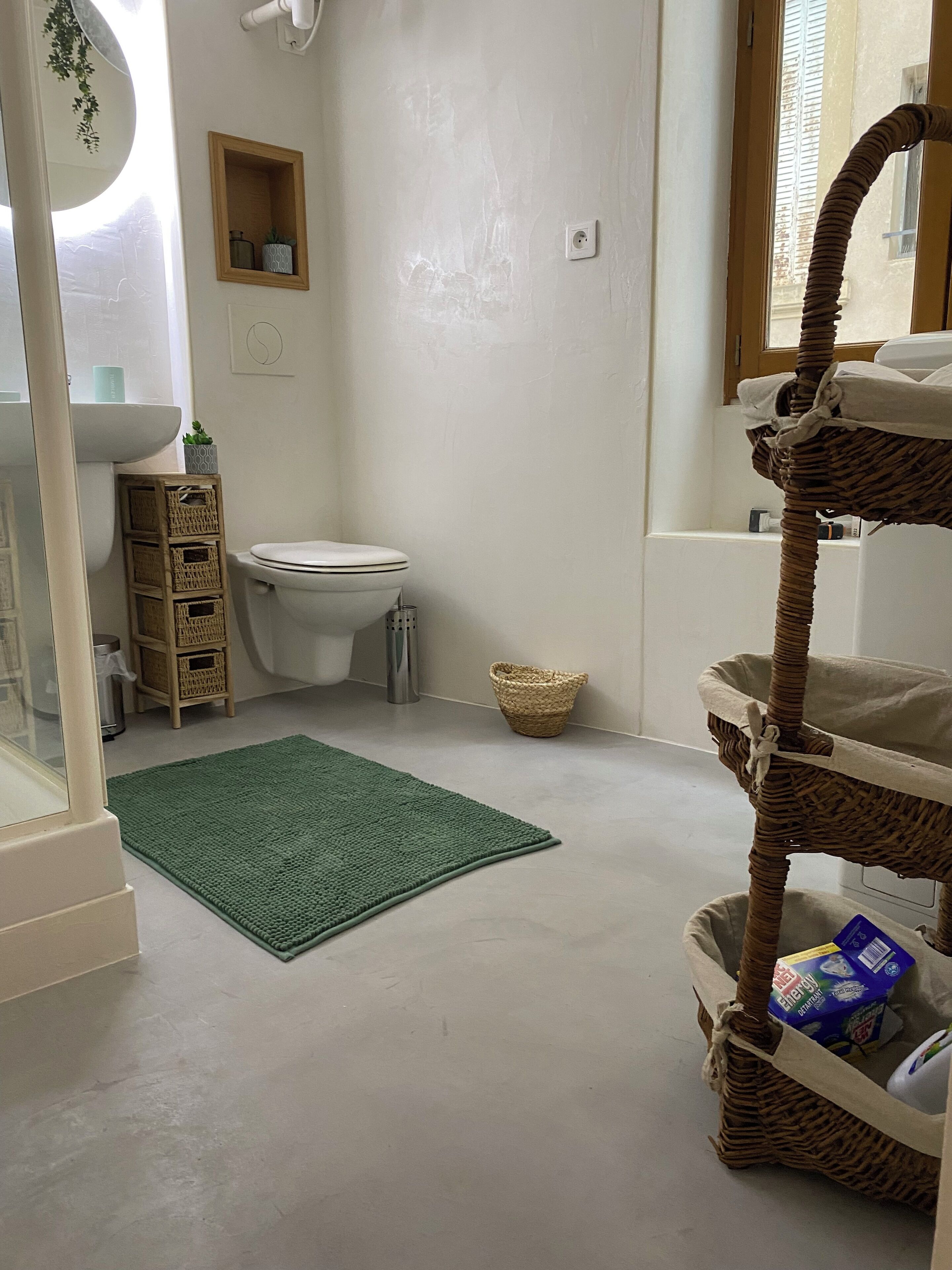 A bathroom with a toilet, a green rug, wicker baskets, and a window.