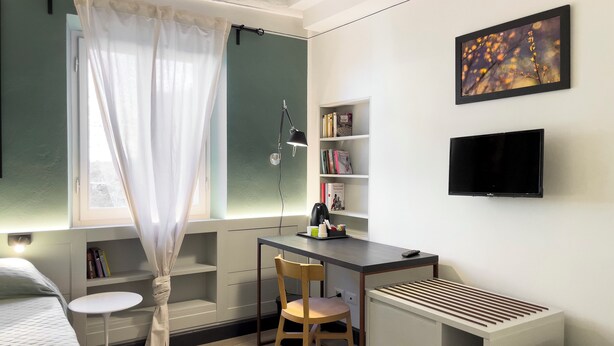 Habitación pequeña con cama, escritorio, silla, estantería, radiador y televisor.