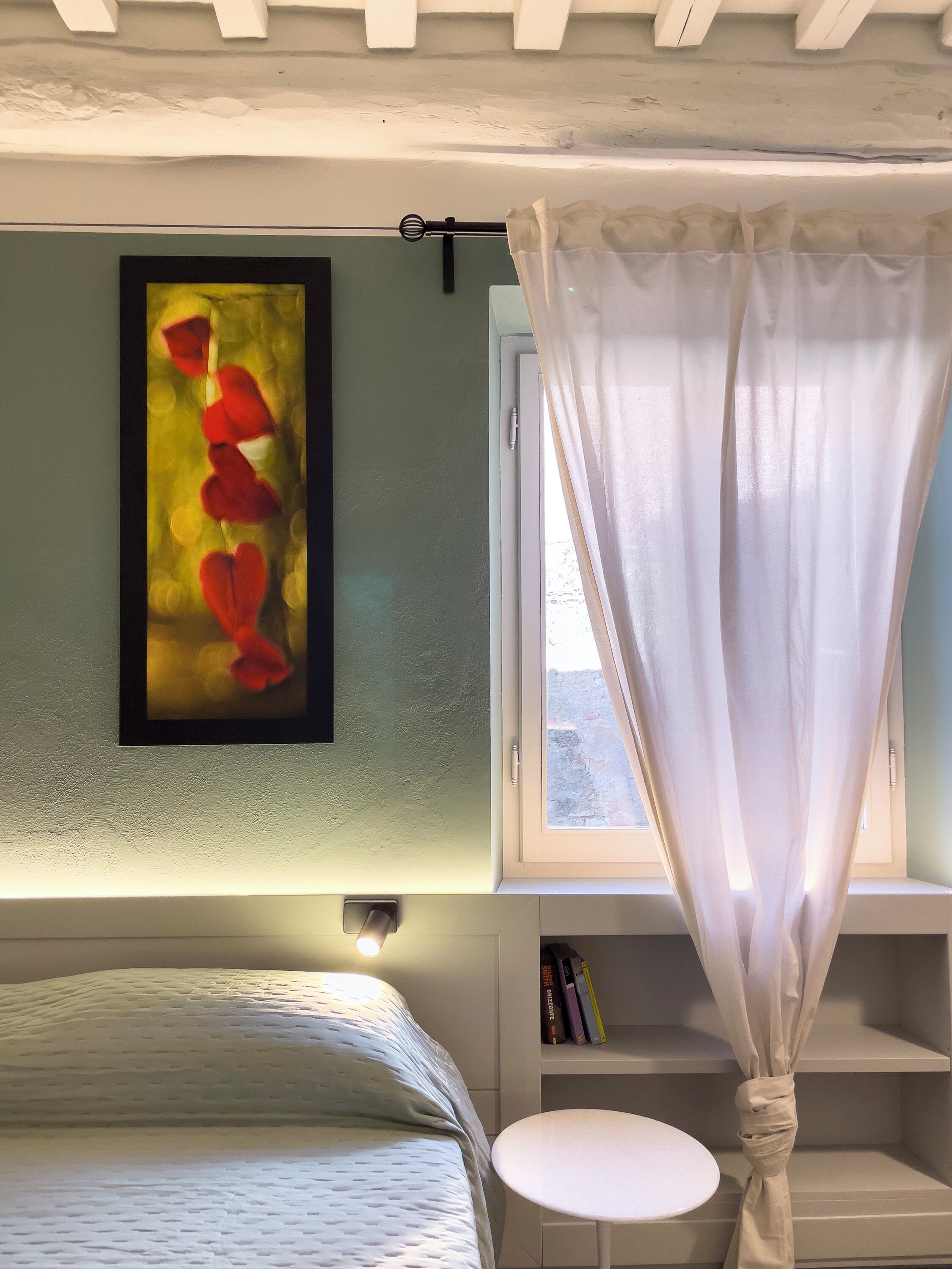 Une chambre à coucher comprenant un lit, une table de chevet, une fenêtre avec des rideaux, une étagère murale avec des livres et une œuvre d’art encadrée.