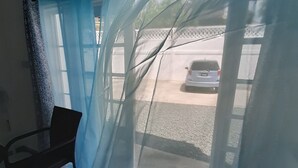 A room with a window covered by a sheer blue curtain, a chair, and a view of a parked car outside. - Luxury Retreat Between City & Rodneybay-free Treat (Castries)