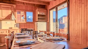 A wooden cabin interior with a dining table set for four, a kitchen area with a stove, and a large window offering a mountain view. - Chalet Altitude - Panoramic view - 8 pers (Bernex)