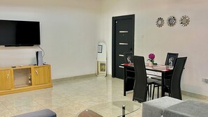 A modern living room with a flat-screen TV, grey sofas, a glass coffee table, and a dining area with a black table and chairs. - ES Homes-2Bedroom Apartment at Achimota (Accra)