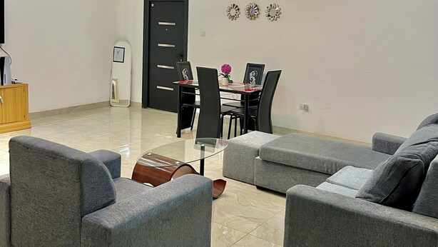A modern living room with grey sofas, a glass coffee table, and a dining area with a black table and chairs. - ES Homes-2Bedroom Apartment at Achimota (Accra)