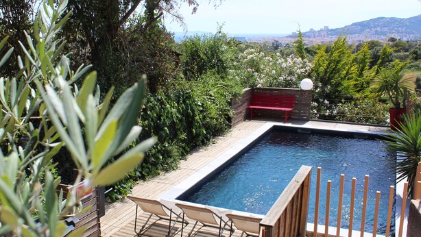 A rectangular swimming pool with a wooden deck, a red bench, and a view of a cityscape in the background.