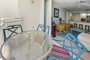 Zona pranzo con tavolo in vetro e sedie blu, cucina con mobili bianchi e soggiorno con divano giallo e tappeto decorato.