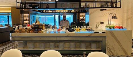 A modern restaurant with a chef at the counter, a bar area, and a seating section with tables and chairs.