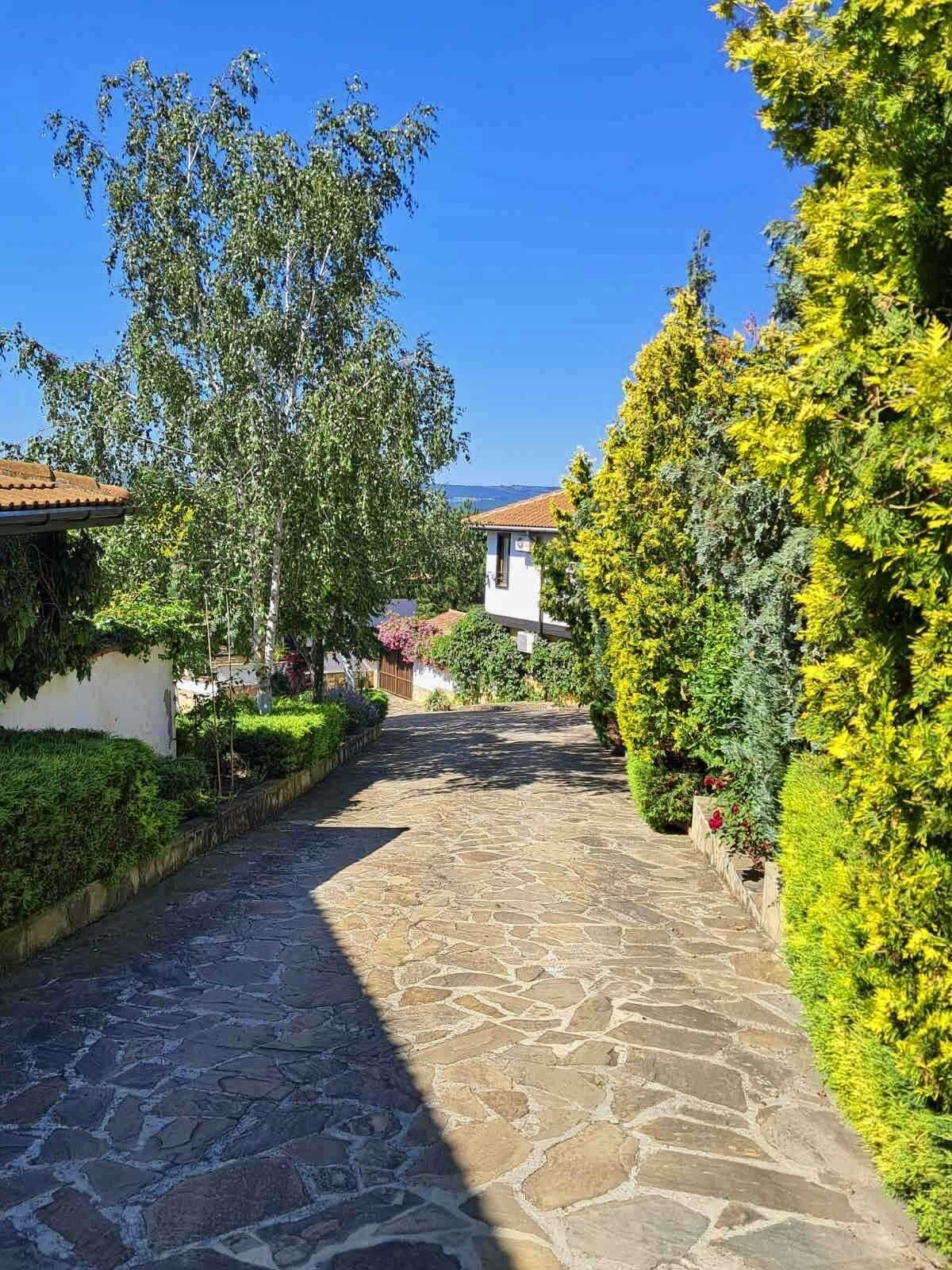 A stone pathway leading to a building with a tiled roof, surrounded by lush greenery and a clear blue sky.