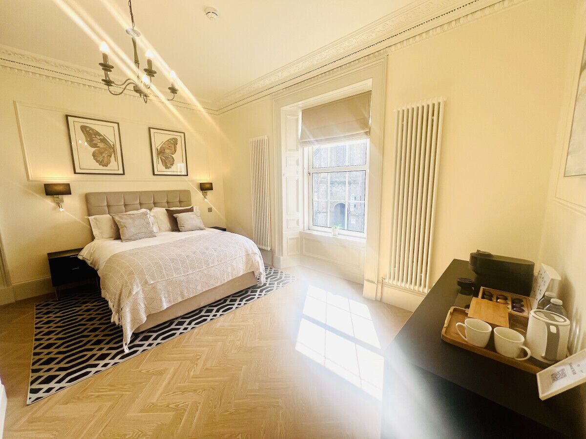 A hotel room with a large bed, a bedside table with a tray of breakfast items, a radiator, and a window with curtains.