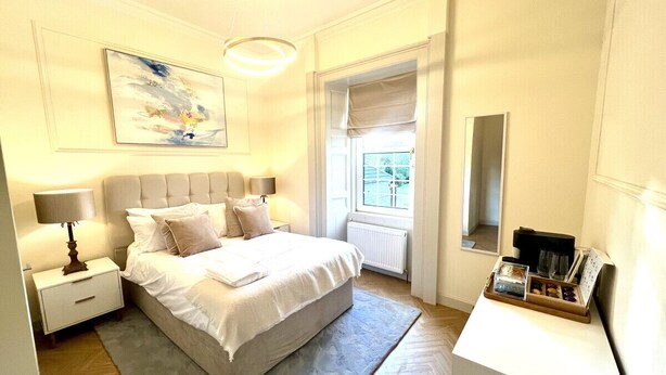 Um quarto com cama, mesas de cabeceira, escrivaninha, janela com cortinas e um quadro na parede.