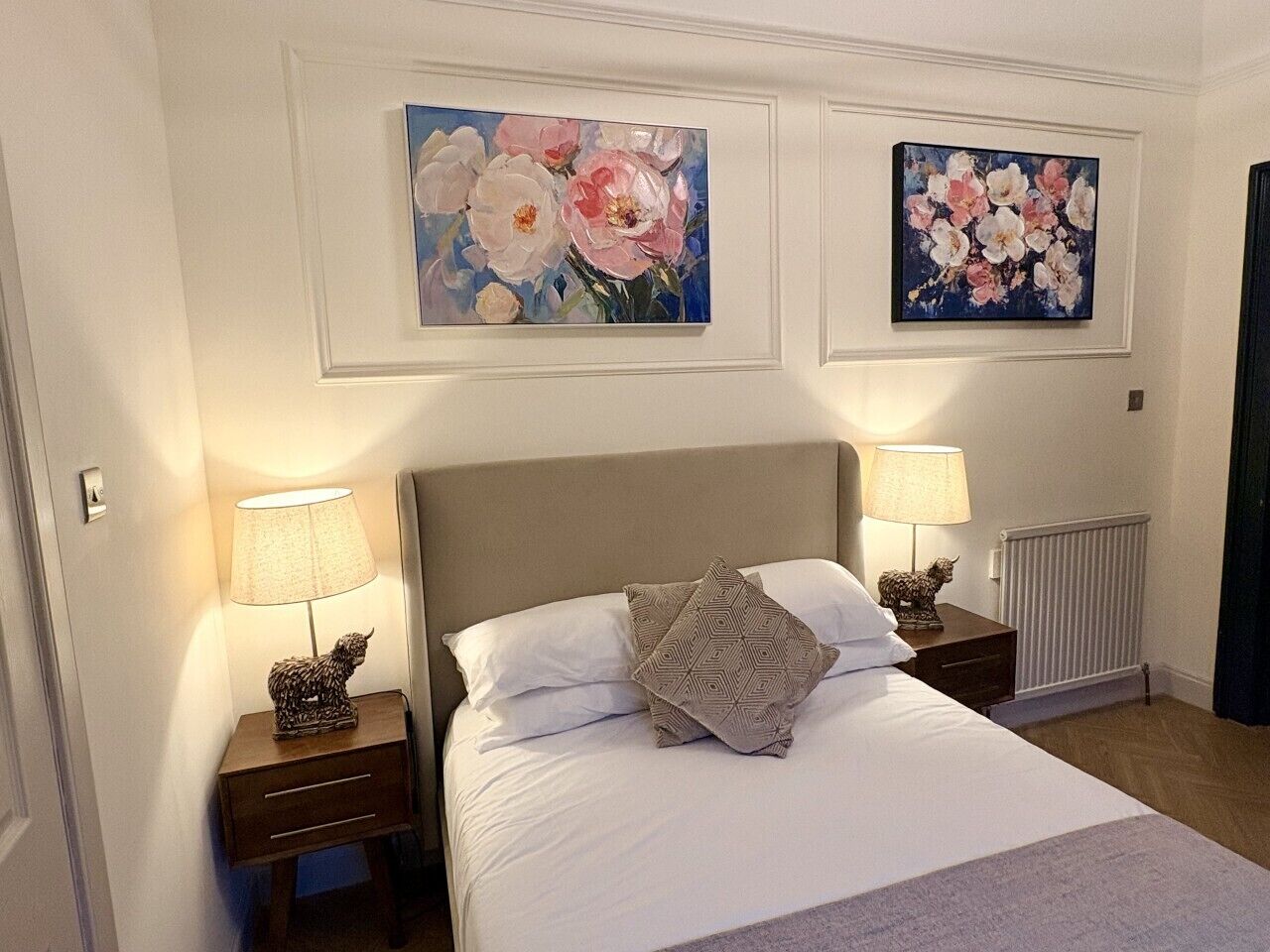 A hotel room with a bed, two nightstands, lamps, a radiator, and two floral paintings on the wall.