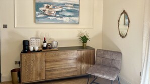 Ein Zimmer mit einem Holzschrank, einem grau gepolsterten Sessel, einer Kaffeemaschine, einer Weinflasche und einem gerahmten Gemälde mit Booten an der Wand.