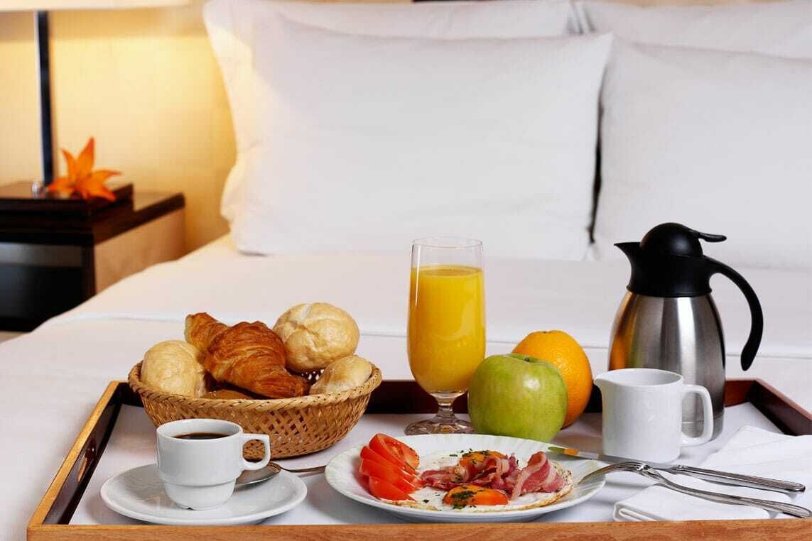 A breakfast tray with croissants, a glass of orange juice, a coffee cup, a plate with eggs and bacon, and a thermos on a bed.