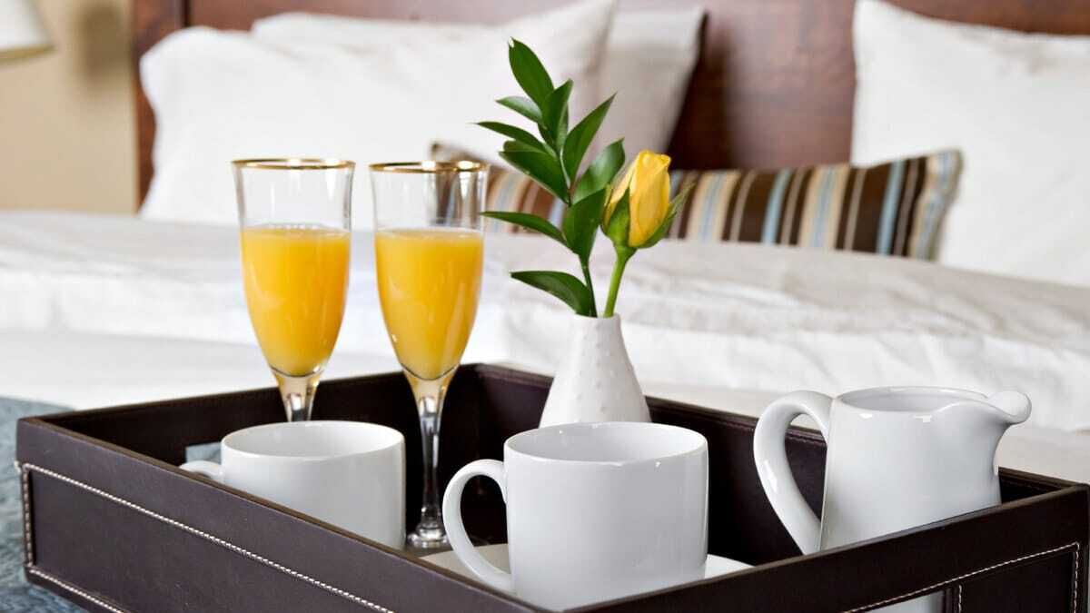 A tray with two glasses of orange juice, a white mug, a white pitcher, and a vase with a yellow flower.