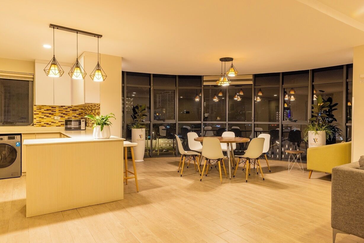 Una cocina moderna con una isla central, zona de comedor con sillas y ventanas amplias con vistas a la ciudad.