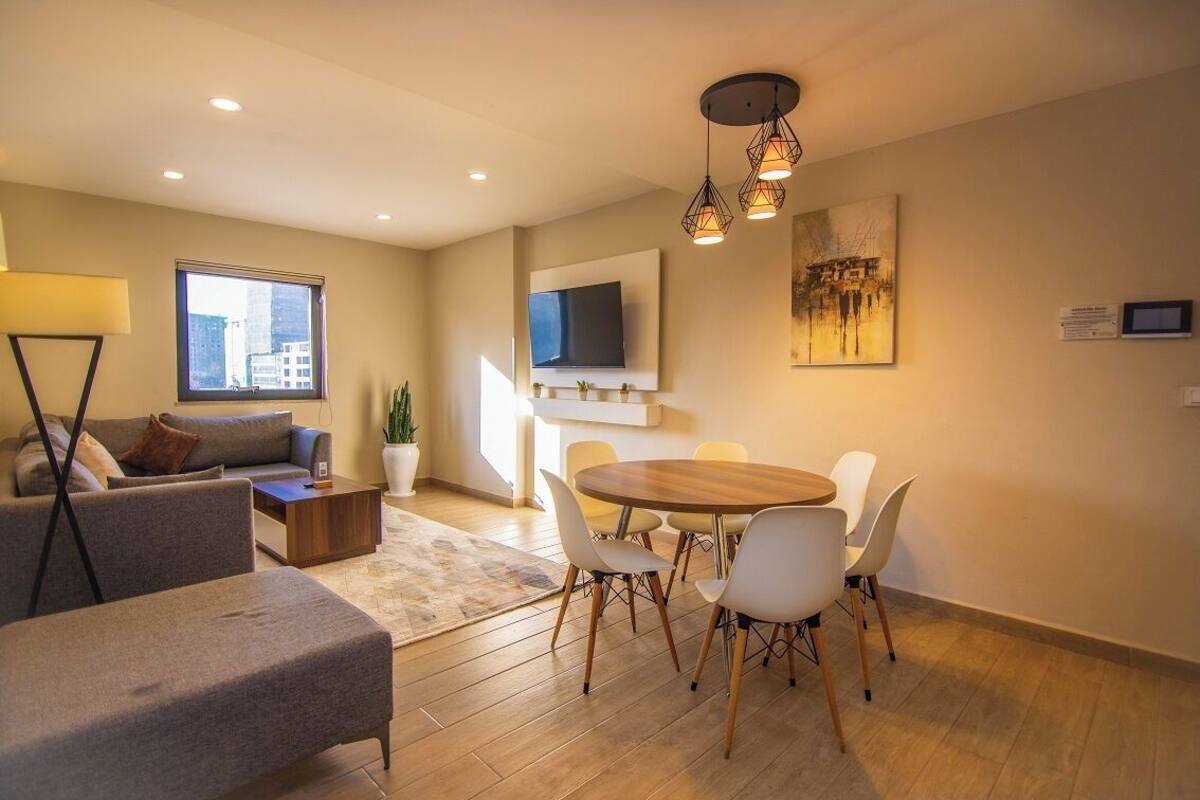 A modern living room with a sofa, a dining table with chairs, a TV mounted on the wall, and a potted plant.