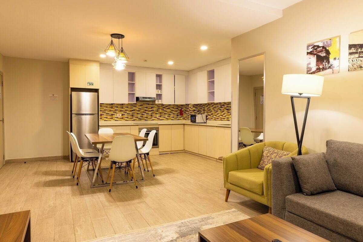 A compact living space with a dining area, a sofa, a coffee table, a kitchen with a refrigerator, and a lamp.