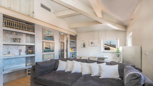 Een moderne woonkamer met een hoekbank, inbouwplanken met decoratieve objecten en uitzicht op de stad door een groot raam.