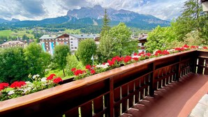 Ein Balkon mit Holzgeländer, der auf eine gebirgige Landschaft blickt, ein Gebäude mit begrüntem Dach und ein Garten mit roten Blüten.
