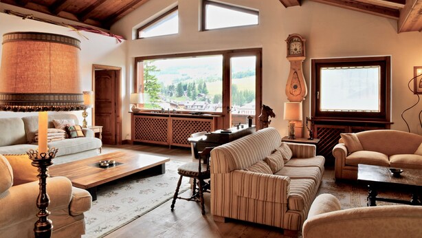 Ein geräumiges Wohnzimmer mit hohen Decken, Holzbalken, einem großen Fenster mit Blick auf die Landschaft, einer traditionellen Uhr und einem gemütlichen Kamin.