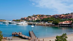 Un port de plaisance situé sur la côte, où sont amarrés des yachts, avec une jetée en bois et des bâtiments aux toits recouverts de tuiles rouges.