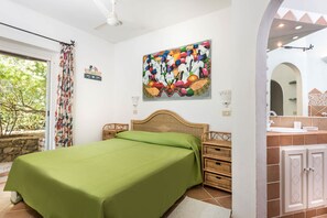 A bedroom with a bed, a painting, a wicker nightstand, a bathroom with a sink and mirror, and a view of trees outside. - NEW! Villa Roberta - walk distance to Porto Cervo (Porto Cervo)