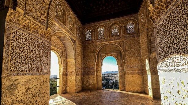 An interior with ornate arches and detailed carvings, a stone floor, and a view through an archway.