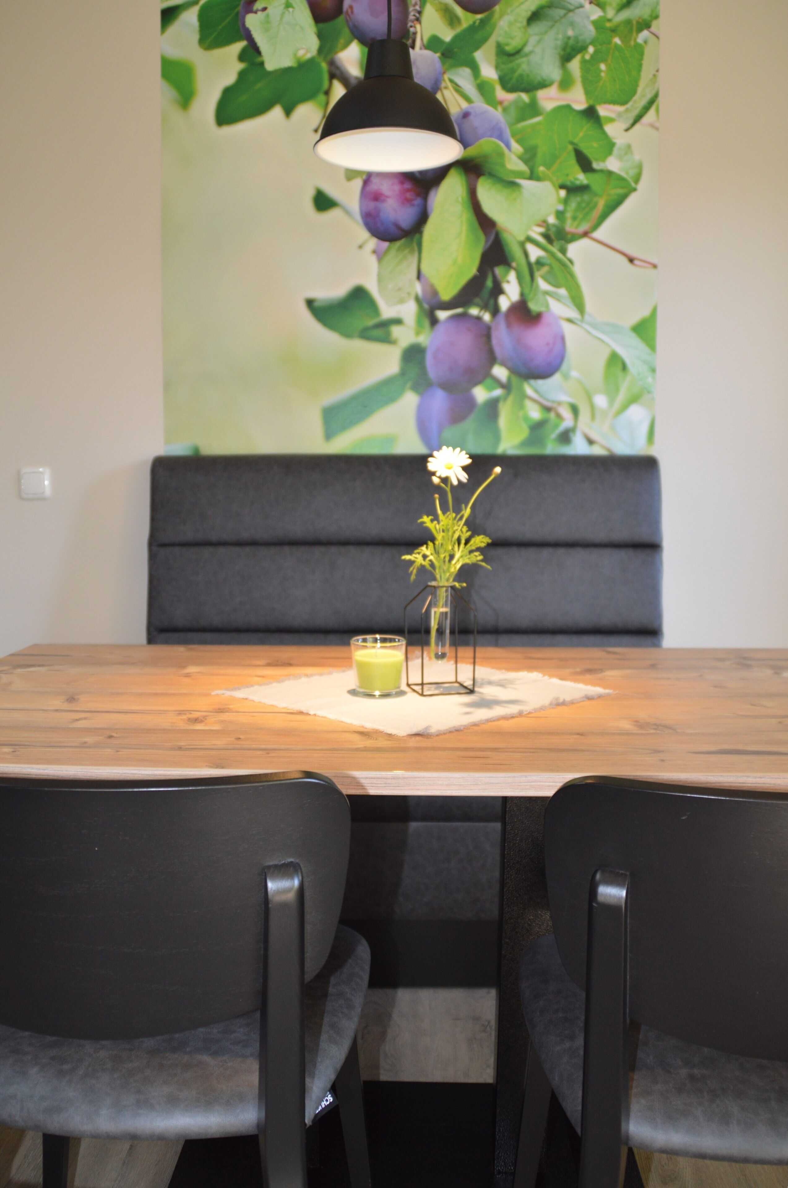 Et spiseområde med et træbord, to stole, en vase med blomster og en vægdekoration med blommer.
