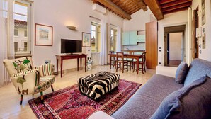 Ein Wohnzimmer mit Sofa, Hocker, Sessel, Fernseher, Esstisch, Stühlen und einer Küchenzeile.
