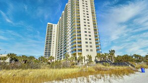 A high-rise hotel with multiple balconies, located near a beach with sandy terrain and sparse vegetation. - Remodeled, open airy condo with full resort access, 1706 Brighton (Myrtle Beach)