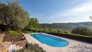 A rectangular swimming pool with a stone deck, surrounded by lush greenery and a view of rolling hills. - Beautiful villa with private garden and pool (Costermano)