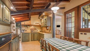 A kitchen with wooden cabinets, a dining table with a checkered tablecloth, and a large window with a view of the interior. - Beautiful villa with private garden and pool (Costermano)