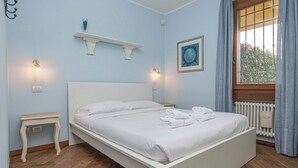 A neatly made bed with white linens and towels, a white nightstand with a lamp, a small shelf with a blue vase, and a window with curtains. - Beautiful villa with private garden and pool (Costermano)