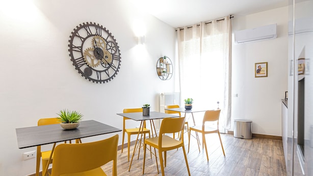 Un comedor con una mesa negra y sillas amarillas, un reloj de pared y una planta en maceta.