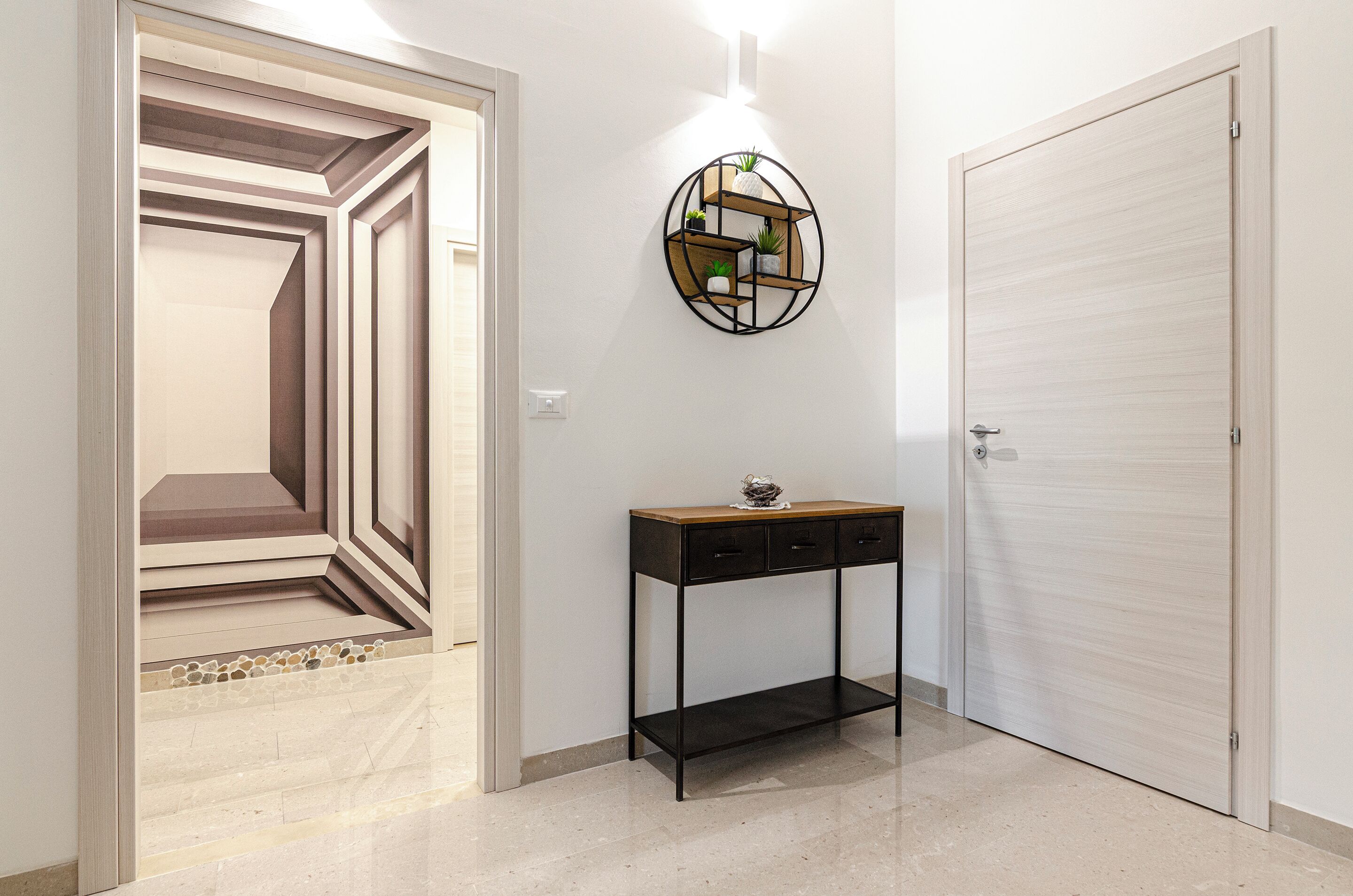 Un pasillo con una puerta de diseño geométrico, un estante circular sujeto a la pared con plantas, y una consola con cajones.
