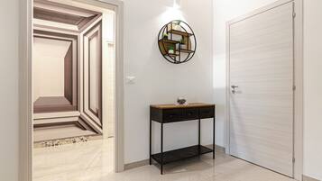 Un pasillo con una puerta de diseño geométrico, un estante circular sujeto a la pared con plantas, y una consola con cajones.
