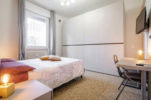 Une chambre à coucher avec un lit, un bureau et une chaise. Il y a une fenêtre avec des rideaux, un radiateur et un téléviseur fixé au mur.